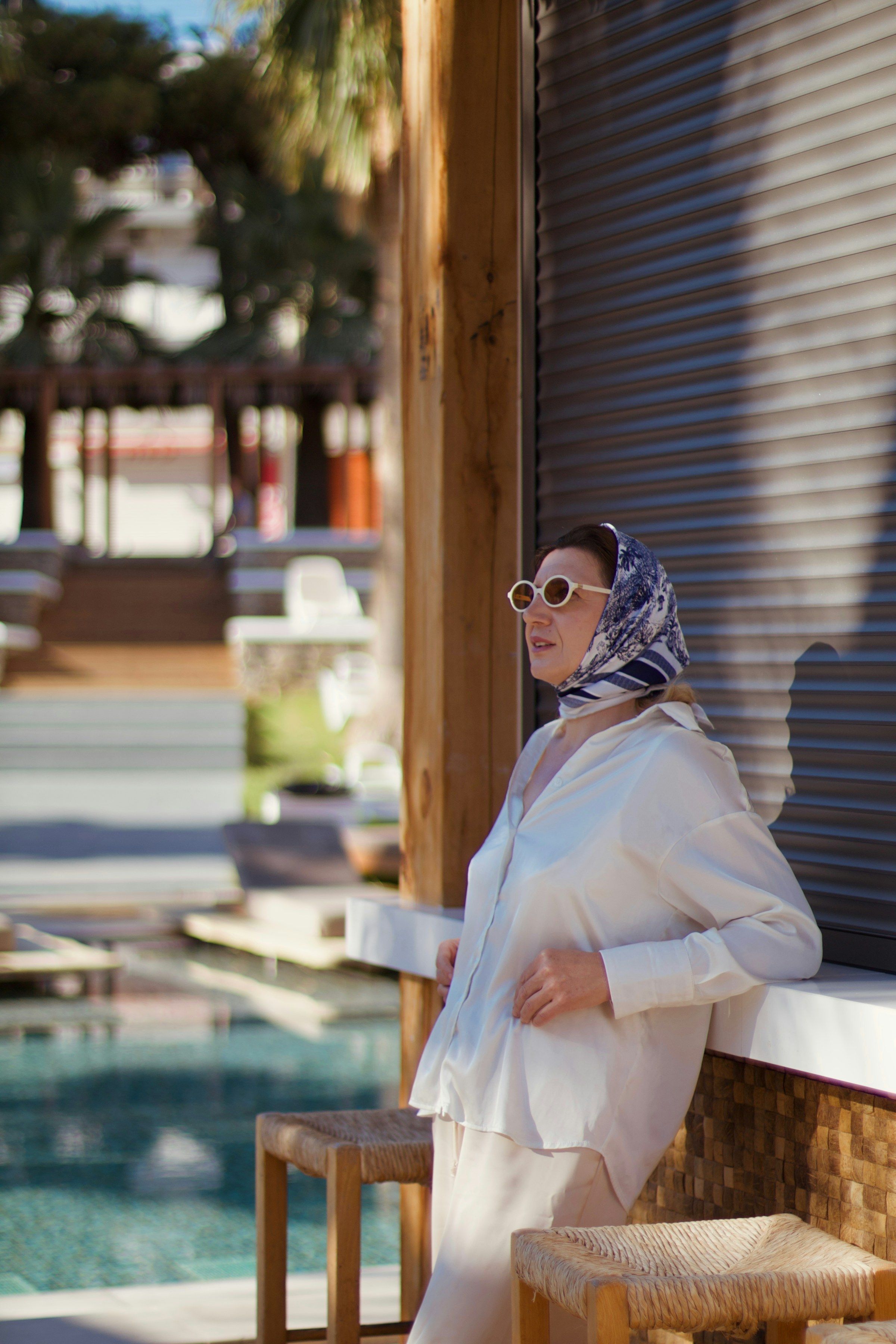 Mulher de lenço estampado, óculos claros e camisa creme encostada numa veneziana, piscina ao fundo